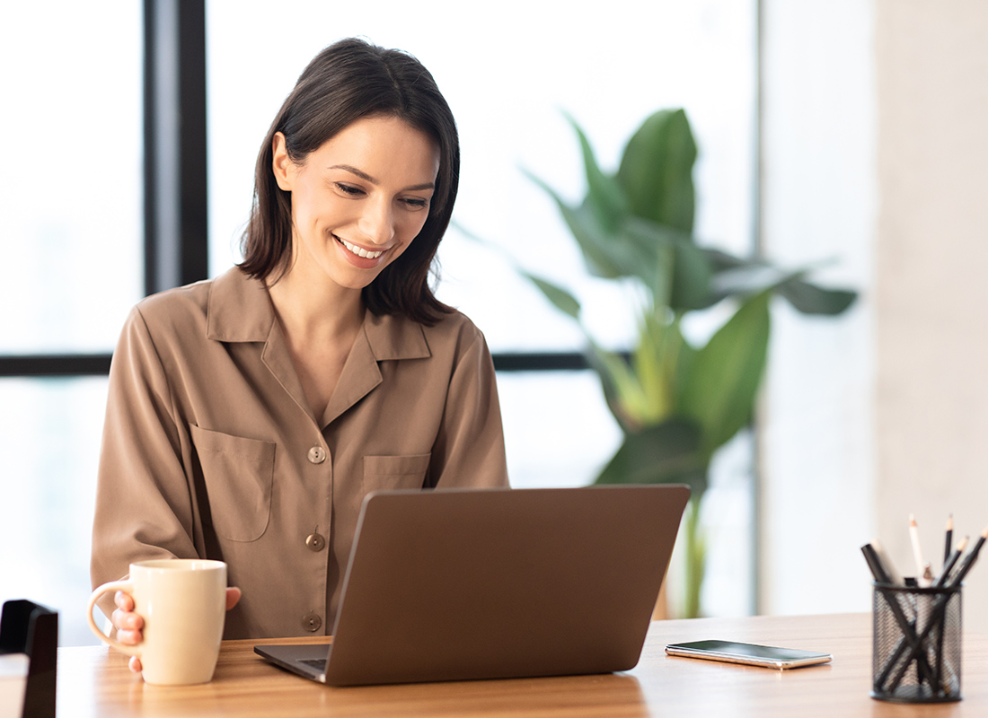 Blog - Woman Sitting and Smiling at Open Laptop on a Wooden Desk While Holding a Mug in Her Modern Home